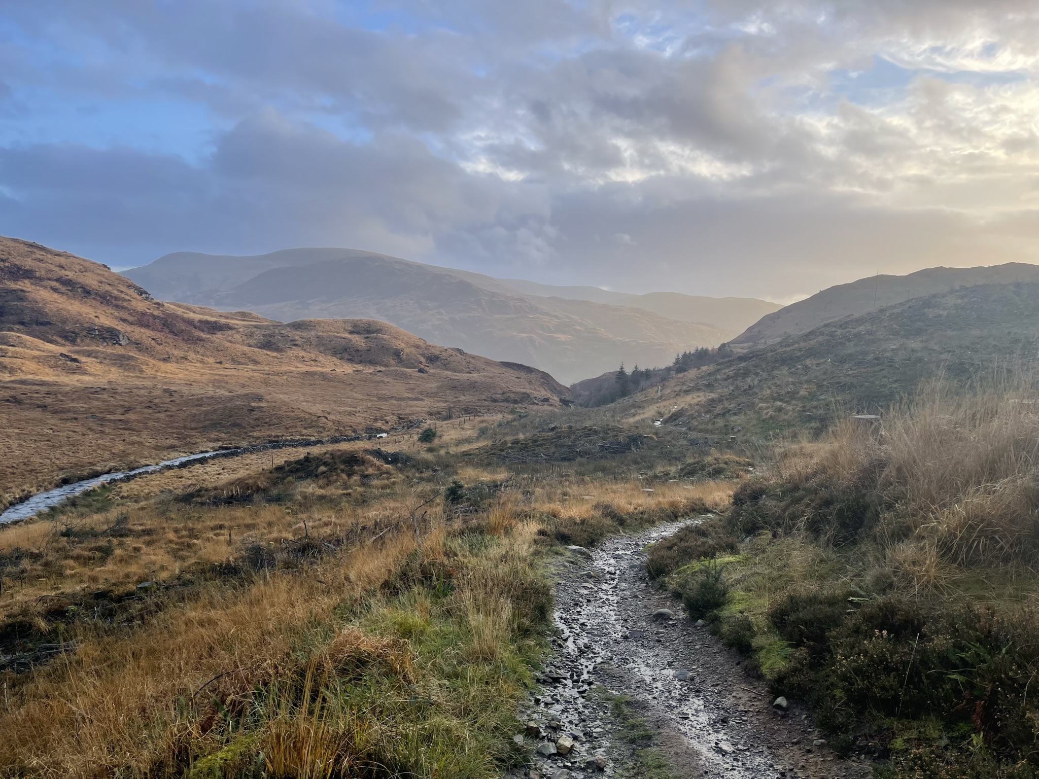 A muddy path winding into a wide mountain valley beneath changing light
