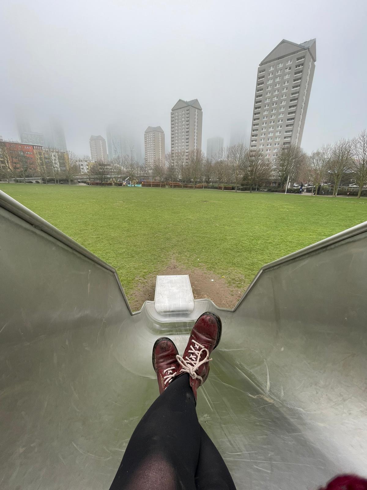 A pair of red boots resting at the top of a metal slide beneath fog-bound tower blocks